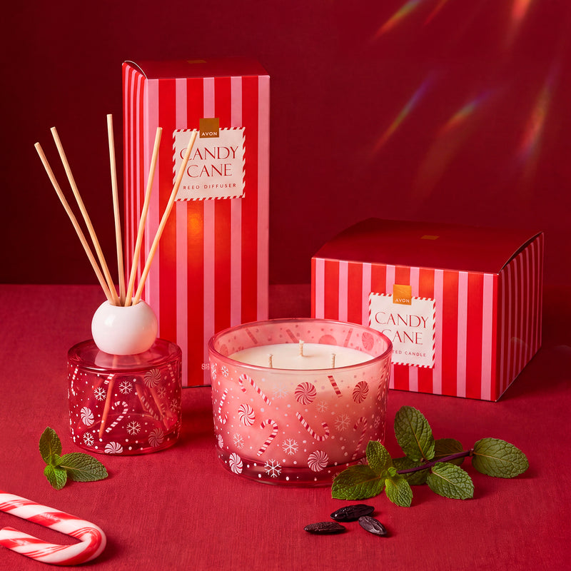 A festive Avon holiday candle and reed diffuser set in red packaging with candy cane and mint props on a red surface against a red background. 