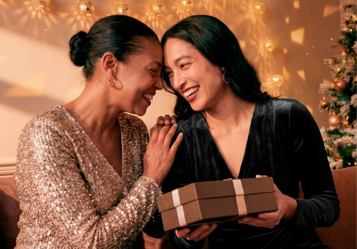Two women smiling and hugging while holding a brown gift box with a white ribbon in a warmly lit holiday setting. One wears a sparkling champagne sequin top, and the other wears a black velvet top.