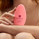 A person holding a pink Avon Her Wisdom Comforting Hand Elixir bottle against their arm and hand, with a softly blurred warm-toned background.