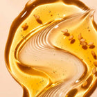 A close-up of Sim Sim Bath & Shower Oil, showing a glossy, translucent golden liquid with scattered small brown seeds on a warm beige background.