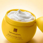 A yellow jar of Avon Her Wisdom whipped oil body cream with white cream visible inside, shown on a warm beige background with the lid resting beside it.