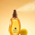 A bottle of Avon Her Wisdom Sim Sim Caring Skin & Hair Perfume Mist with a brown spray nozzle, shown spraying a fine mist against a warm golden-yellow background with a yellow sphere prop.